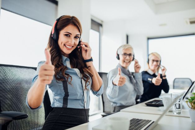 Kundenservicebild, mehrere Damen mit Headset schauen lächelnd mit Daumen hoch nach rechts zum Zuschauer